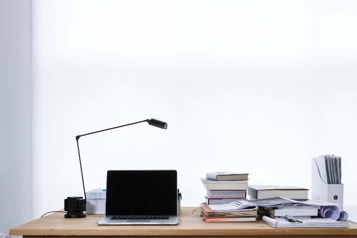 A table with a laptop, lamp, books, and stacks of papers on top.